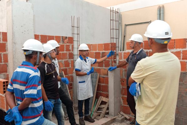 Foto colorida de vários homens com capacete em um espaço de obra durante o curso de capacitação de pedreiro - Metrópoles 