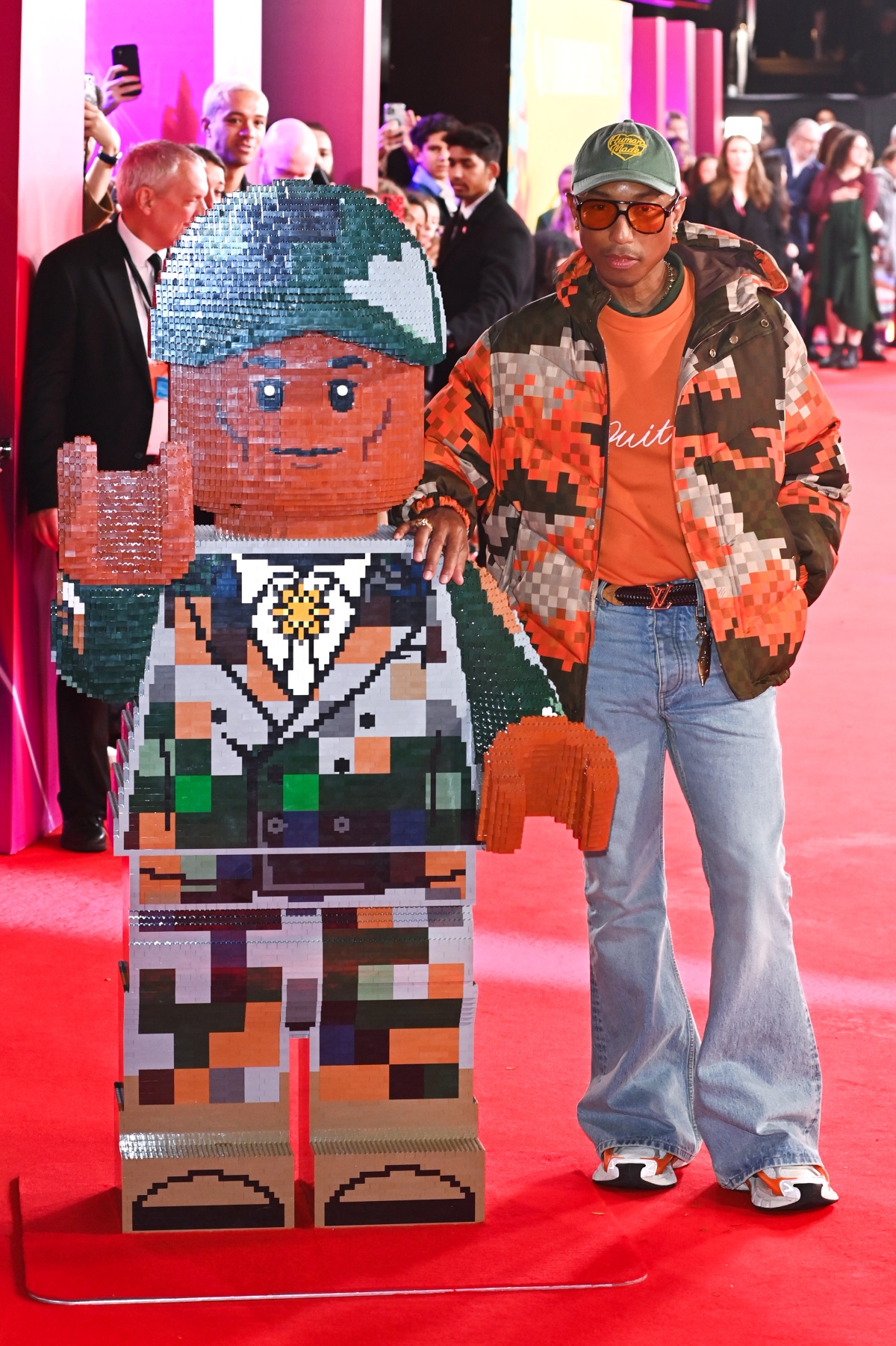 O artista Pharrell Williams posa para a foto. Em um tapete vermelho, ele para ao lado de uma escultura de Lego que recria seu próprio visual